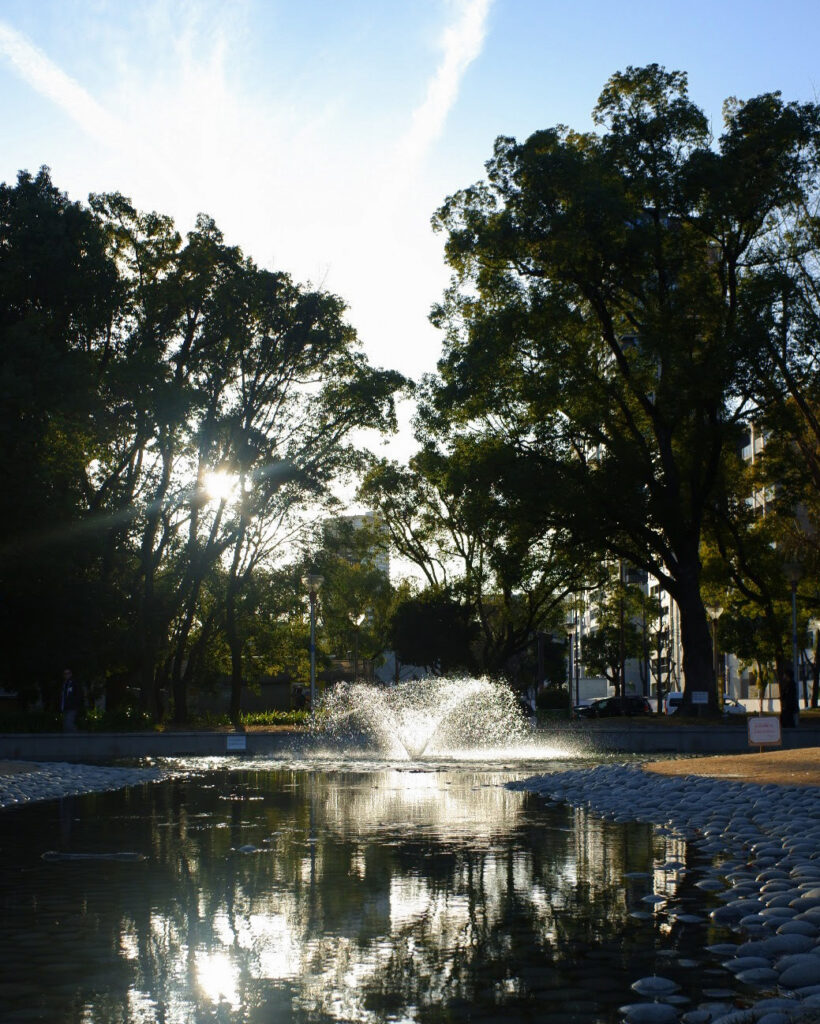 靭公園の中にある噴水の写真。噴水は光を浴びて光っているが、噴水の後ろの木は逆光気味で黒く写っている。