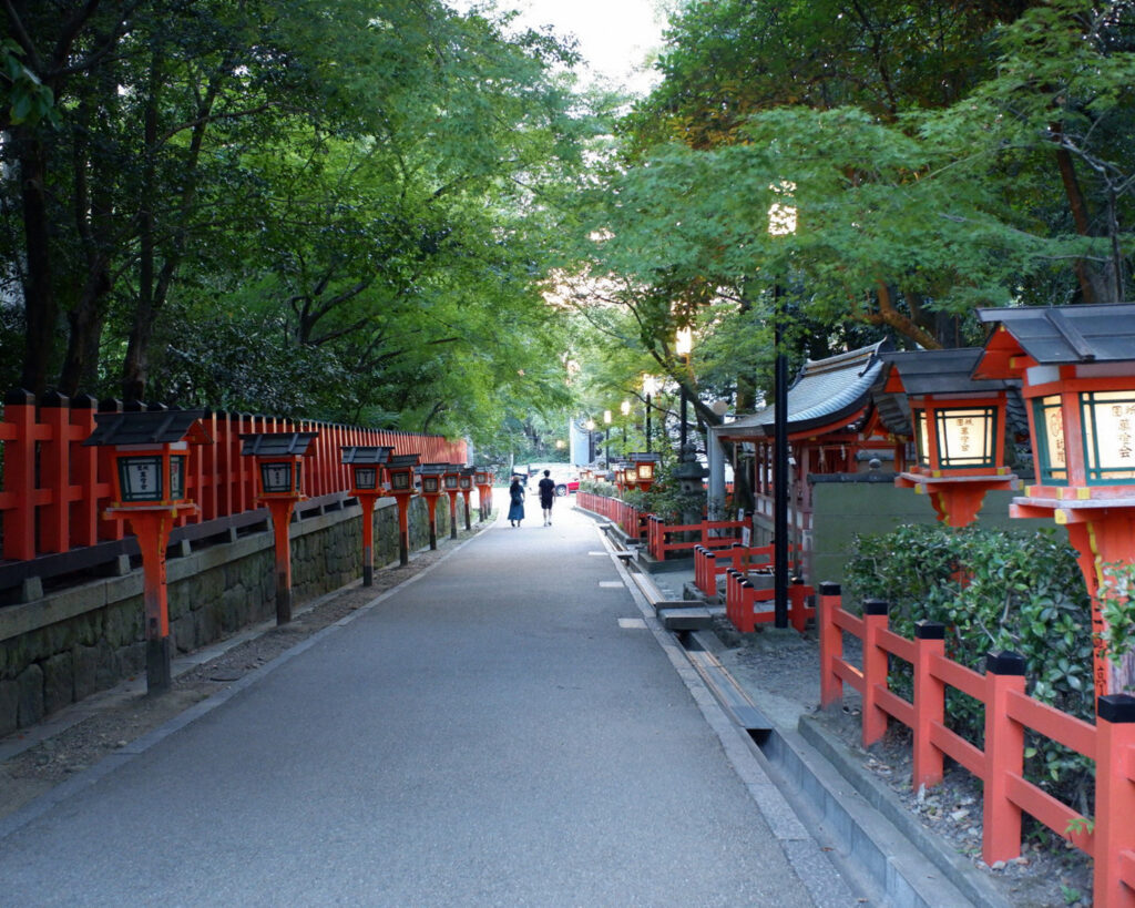 昼の八坂神社の境内。真っ直ぐの道の脇に灯籠や柵がある。その上には木の緑があり、日に照らされている。