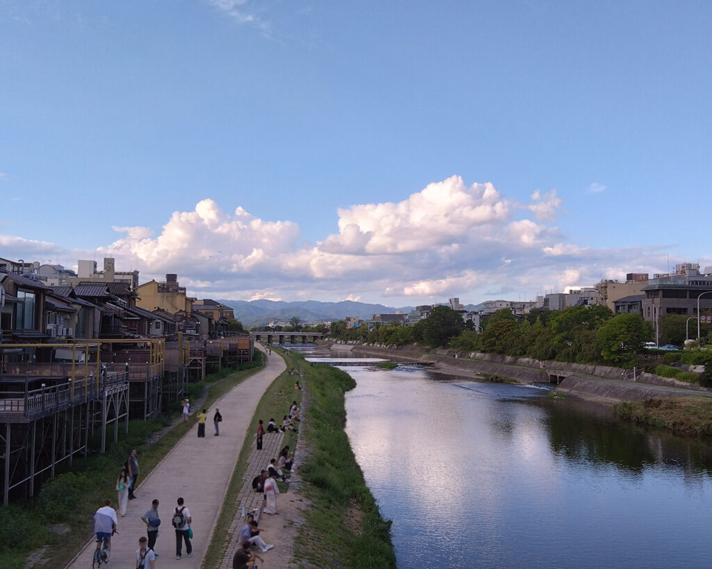 遠くの空に雲がある青空。川にそれが反射している。河川敷は人が歩いたり座ったりしている。